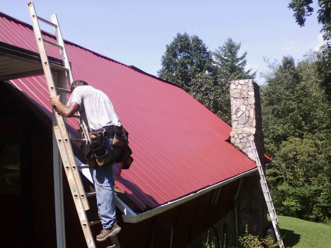 Roofer on a ladder beside a red metal roof and stone chimney — Soffit & Fascia Repair by Roofing Brothers in Kirkwood